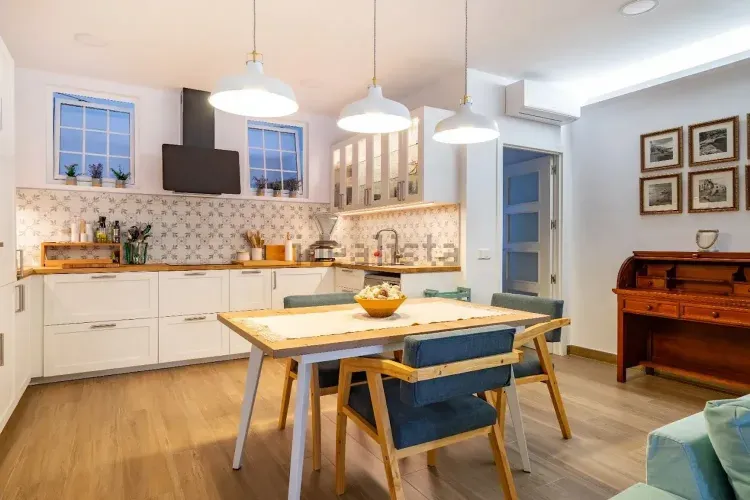 Cocina y comedor modernos y luminosos con armarios blancos, mesa de madera, sillas azules y lámparas colgantes.