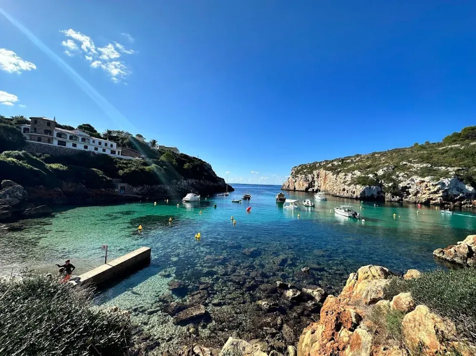 Cala rocosa junto al mar con aguas cristalinas, bañistas y un muelle de madera bajo un cielo brillante.