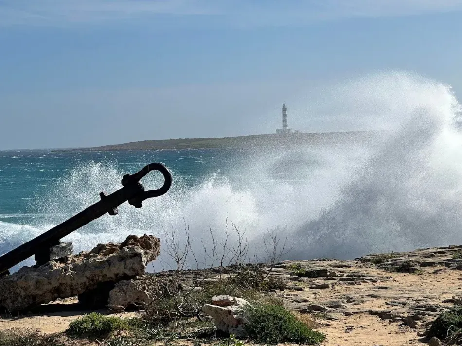 Olas rompiendo contra una costa rocosa, con un ancla negra en primer plano y un faro a lo lejos.