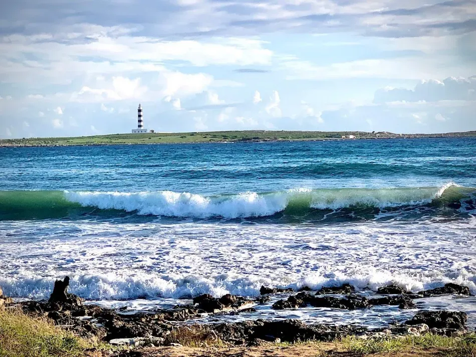 Olas rompiendo en una costa rocosa con un faro a lo lejos bajo un cielo azul brillante.