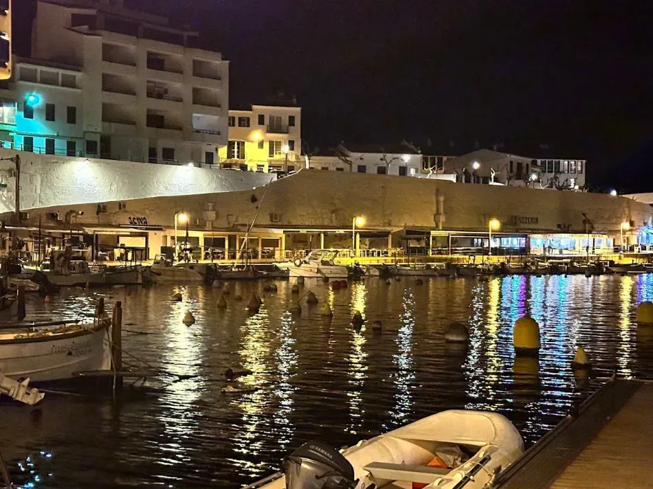 Puerto nocturno con edificios iluminados a orillas del agua y barcos que se reflejan en las aguas tranquilas.