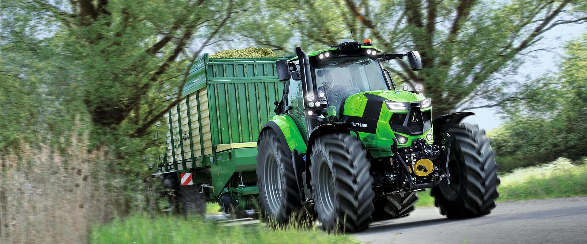 Ein grüner Traktor zieht einen Anhänger auf einer von Bäumen gesäumten Landstraße.