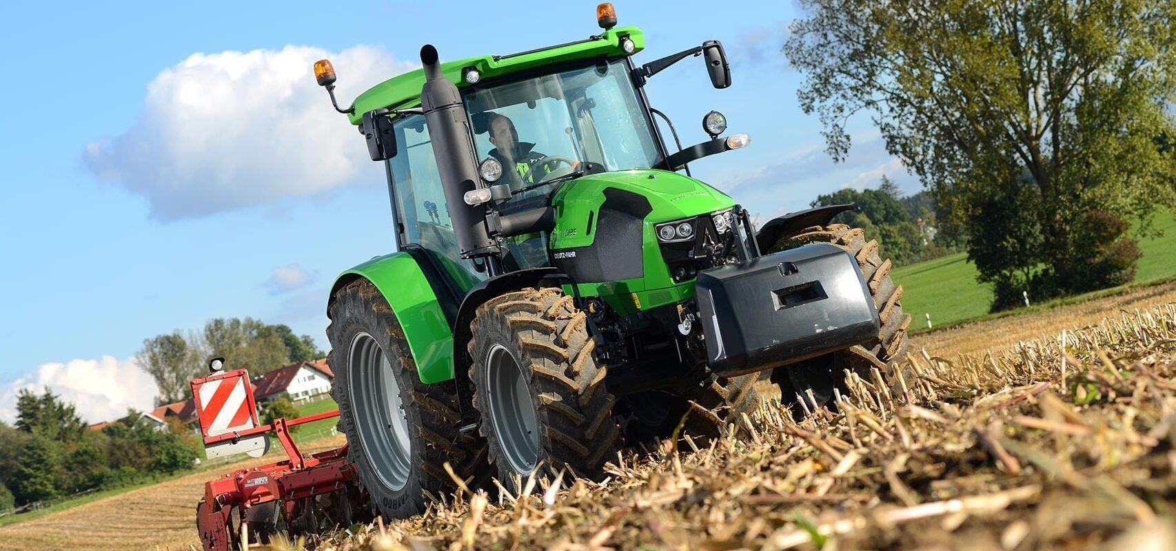 Ein grüner Traktor pflügt an einem sonnigen Tag ein Feld; im Hintergrund ist eine weitere landwirtschaftliche Maschine zu sehen.