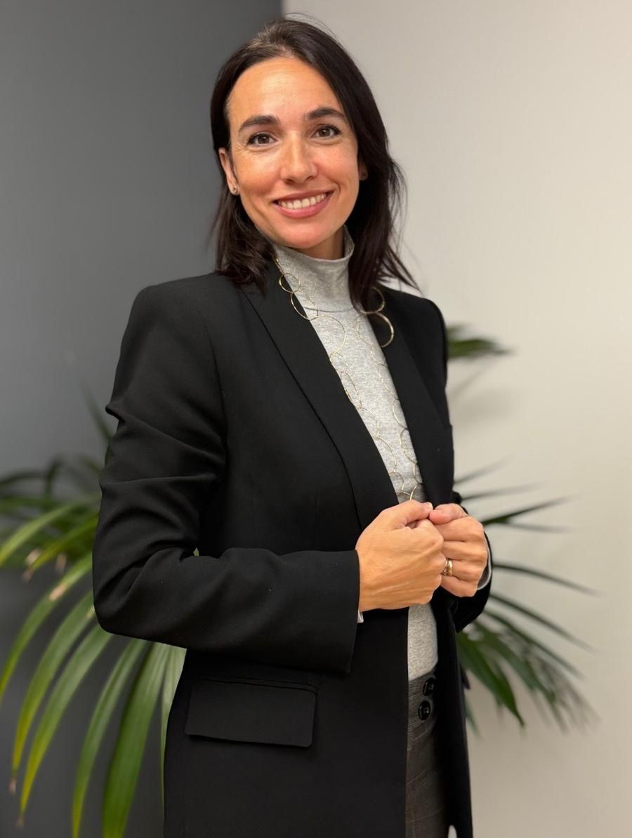 Mujer con blazer negro y jersey de cuello alto gris, sonriendo con las manos entrelazadas.