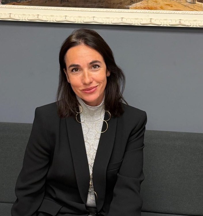 Una mujer con blazer negro y jersey de cuello alto gris sonríe, sentada contra una pared gris.