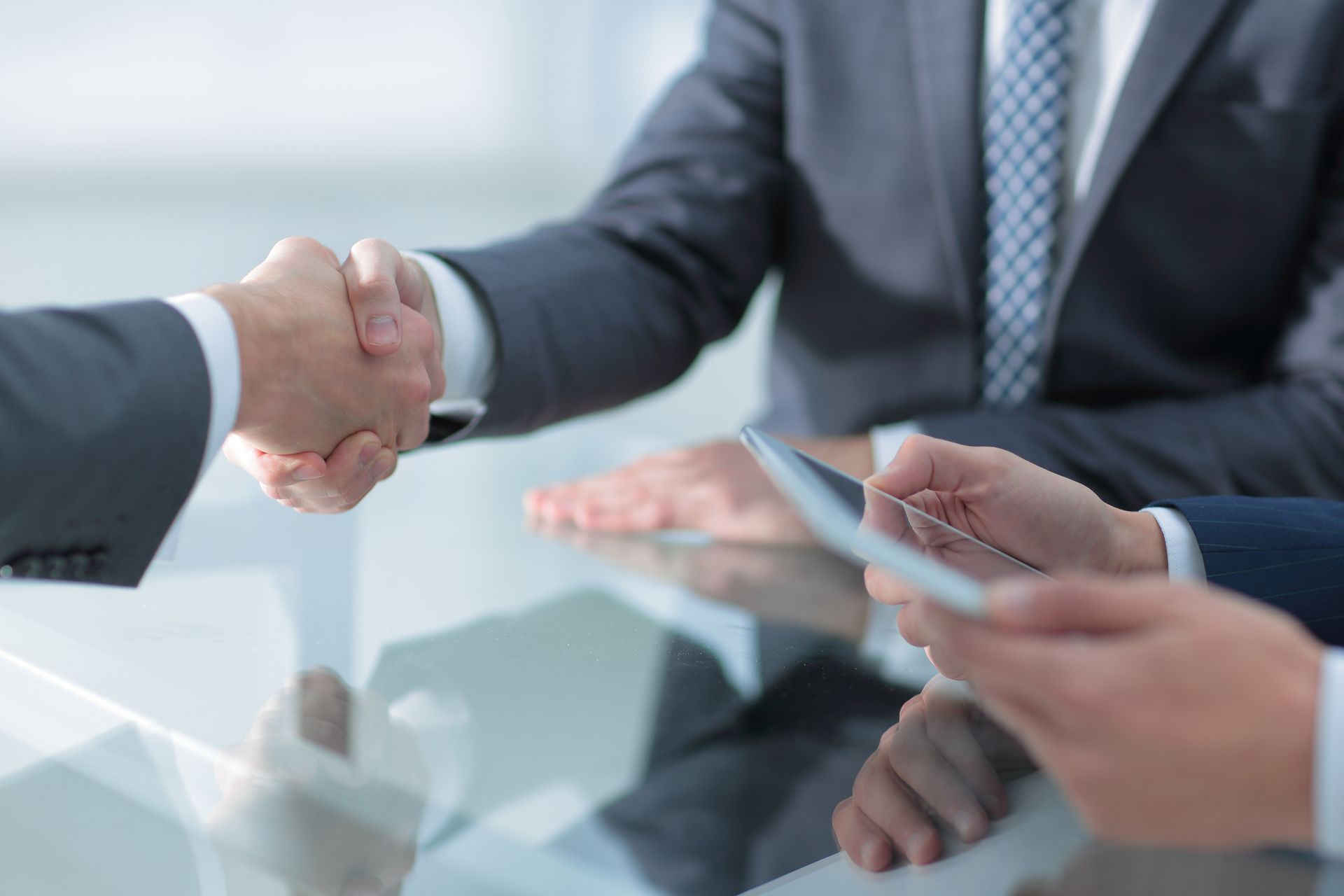 Dos empresarios con traje se dan la mano en una mesa; otra persona sostiene una tableta.