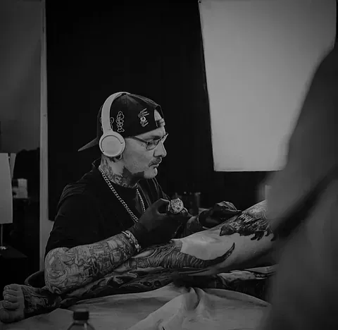 Un tatuador, con auriculares y gorra de béisbol, trabaja en un gran tatuaje en la pierna de un cliente en un estudio.