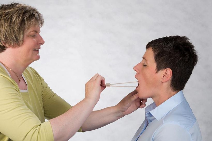 Der Untersucher untersucht mit einem Zungenspatel den Mund eines sitzenden Patienten.