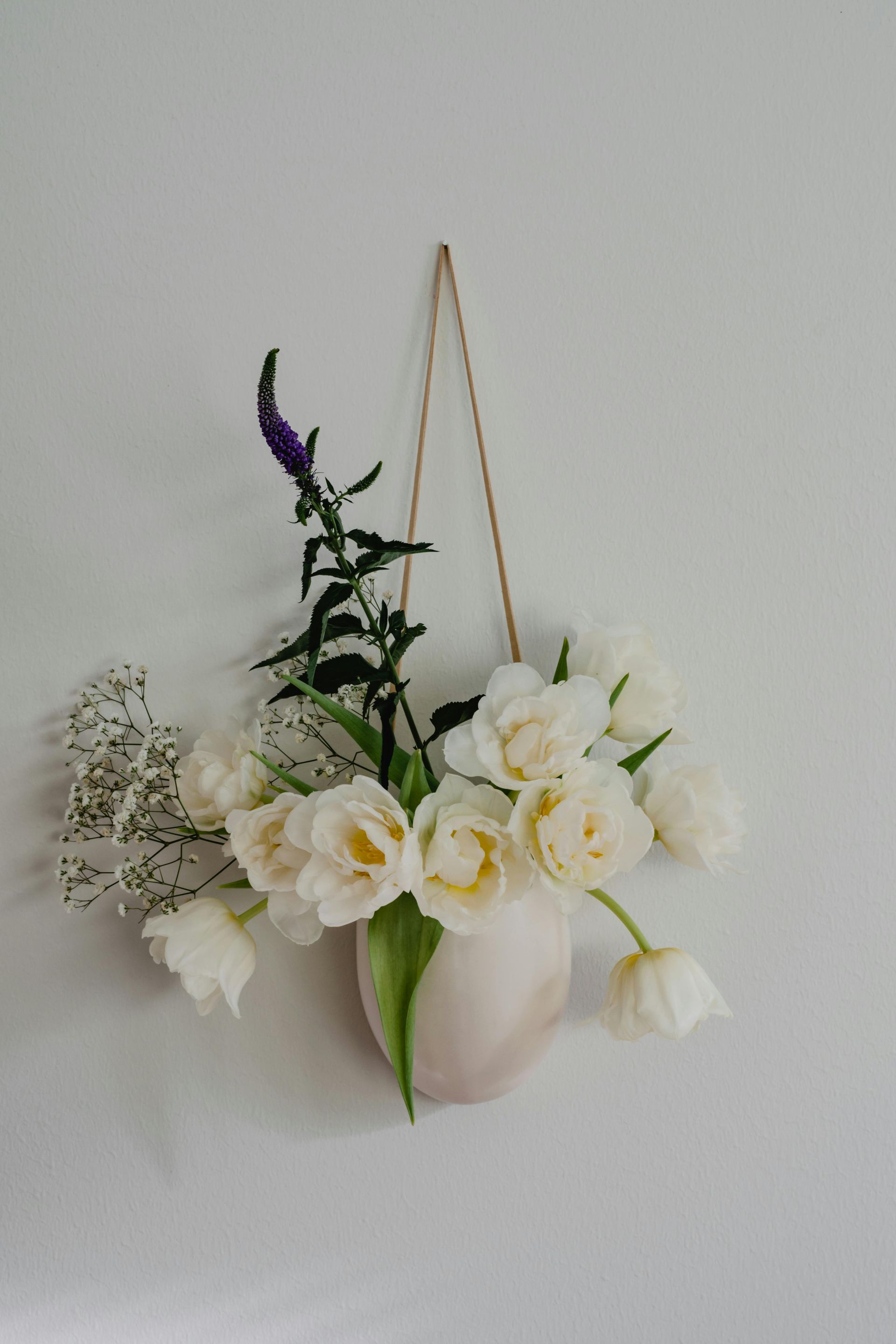 An einer weißen Wand hängt eine Vase mit weißen Blumen.
