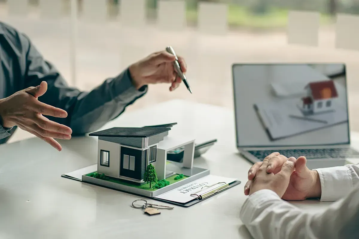 Immobilienmakler präsentiert einem Kunden ein Hausmodell und einen Laptop.