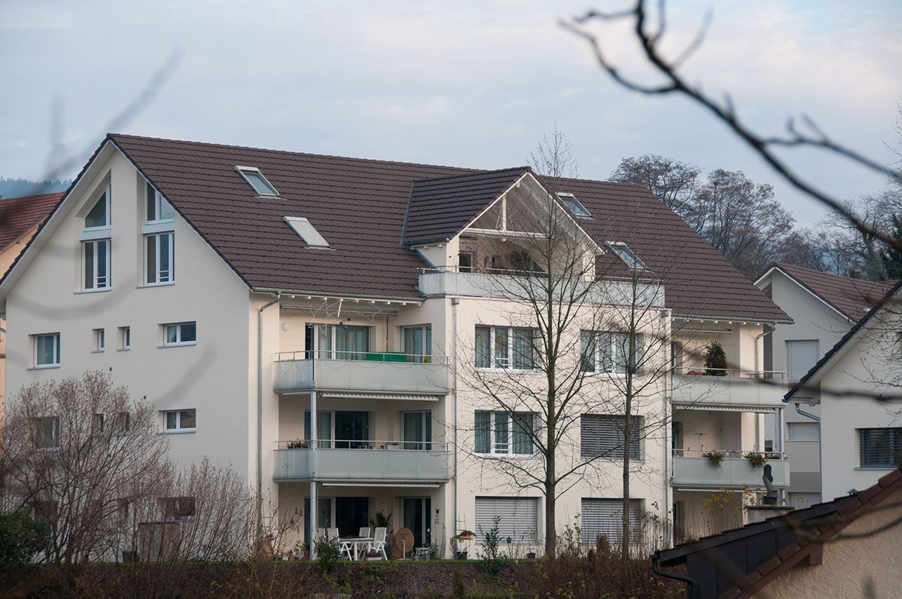 Weißes Wohnhaus mit braunem Dach, Balkonen und Bäumen davor.
