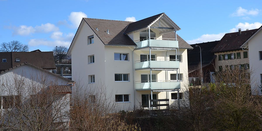 Weißes Mehrfamilienhaus mit Balkonen, braunem Dach und blauem Himmel.