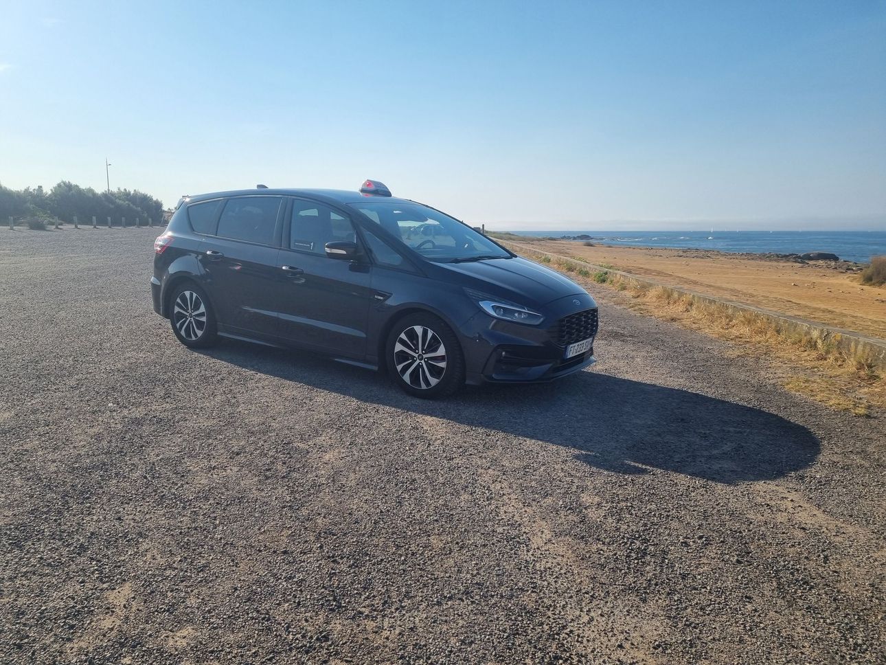 Un SUV sombre garé sur un chemin de gravier au bord de la côte, sous un ciel bleu azur.
