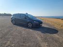 Voiture sombre garée sur un chemin de gravier en bord de mer, sous un ciel bleu azur.