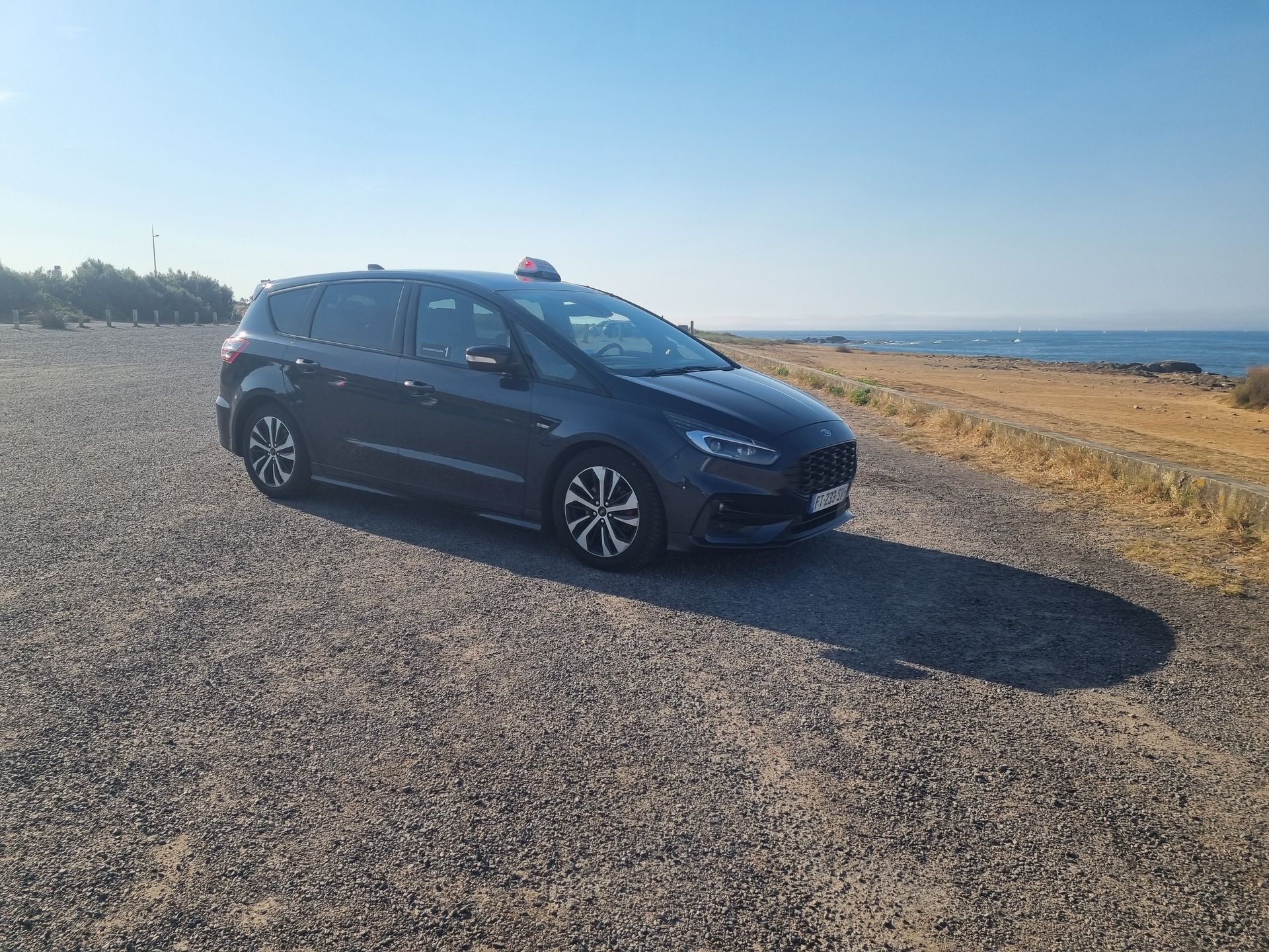 Un SUV sombre garé sur un chemin de gravier au bord de la côte, sous un ciel bleu azur.
