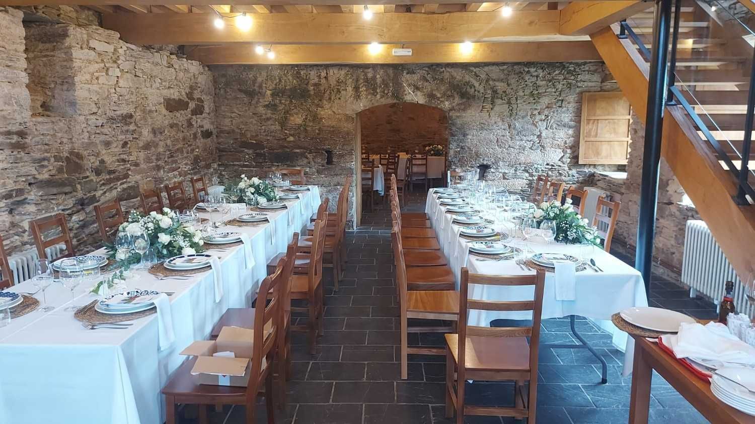 Mesas largas dispuestas para un banquete en un salón rústico con paredes de piedra, tonos neutros e iluminación suave.