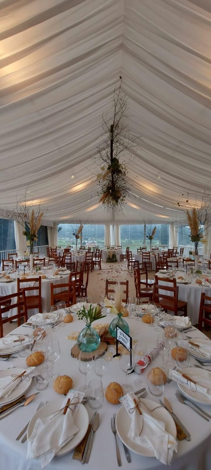 Montaje de recepción de boda: mesas redondas con manteles blancos, sillas de madera.