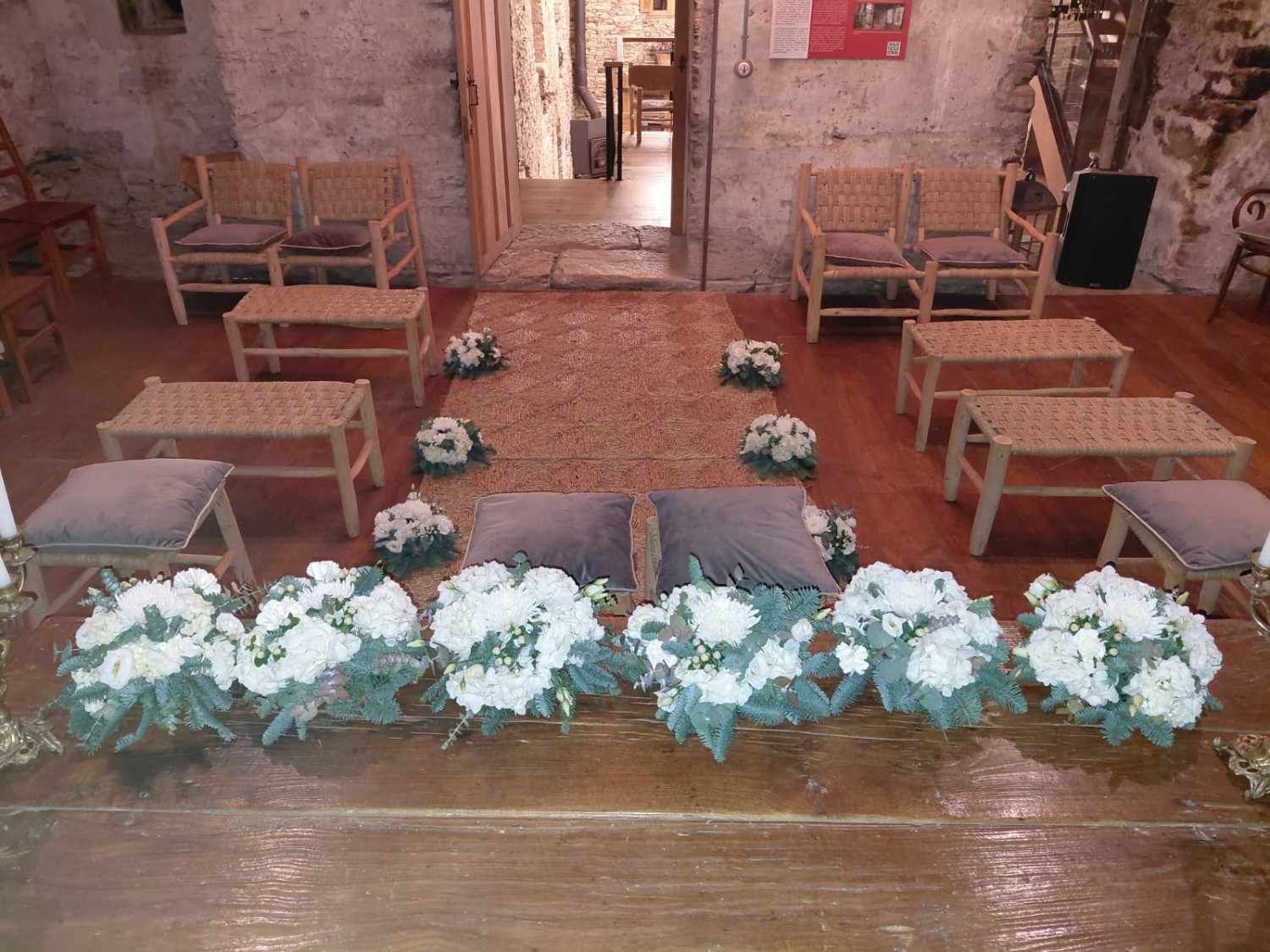 Montaje de la ceremonia de boda: bancos de madera, arreglos florales y almohadas sobre una plataforma elevada.