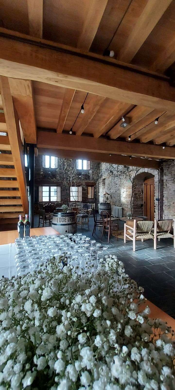 Interior de un edificio con paredes de piedra y vigas de madera, flores en primer plano y una escalera a la izquierda.