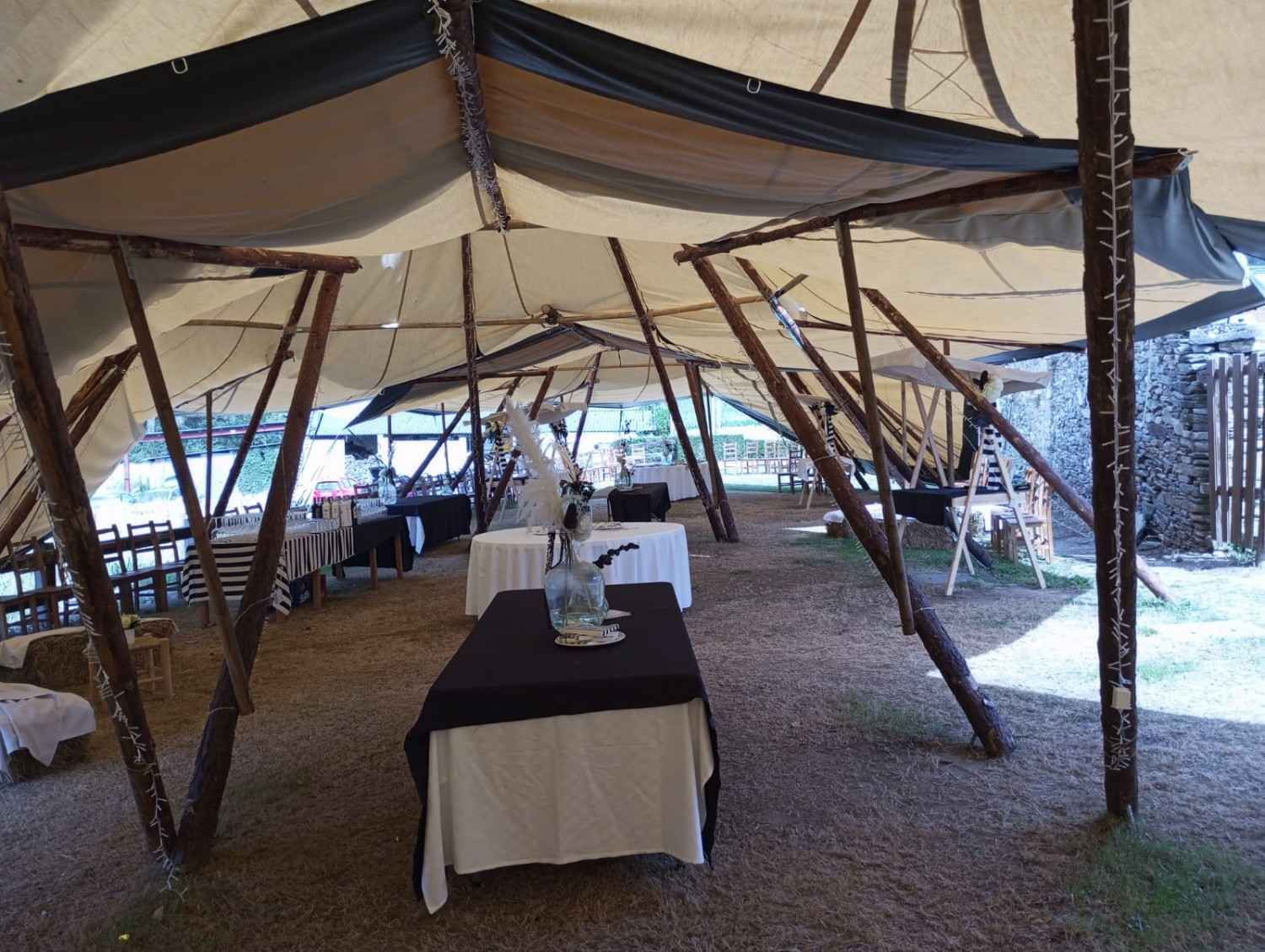 Carpa con mesas preparadas para fiesta, soportes de madera y techo de lona en un entorno exterior con césped.