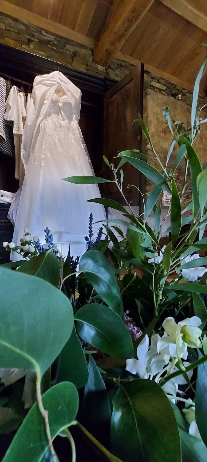 Vestido de novia colgado en un armario, parcialmente oculto por follaje verde y flores blancas.