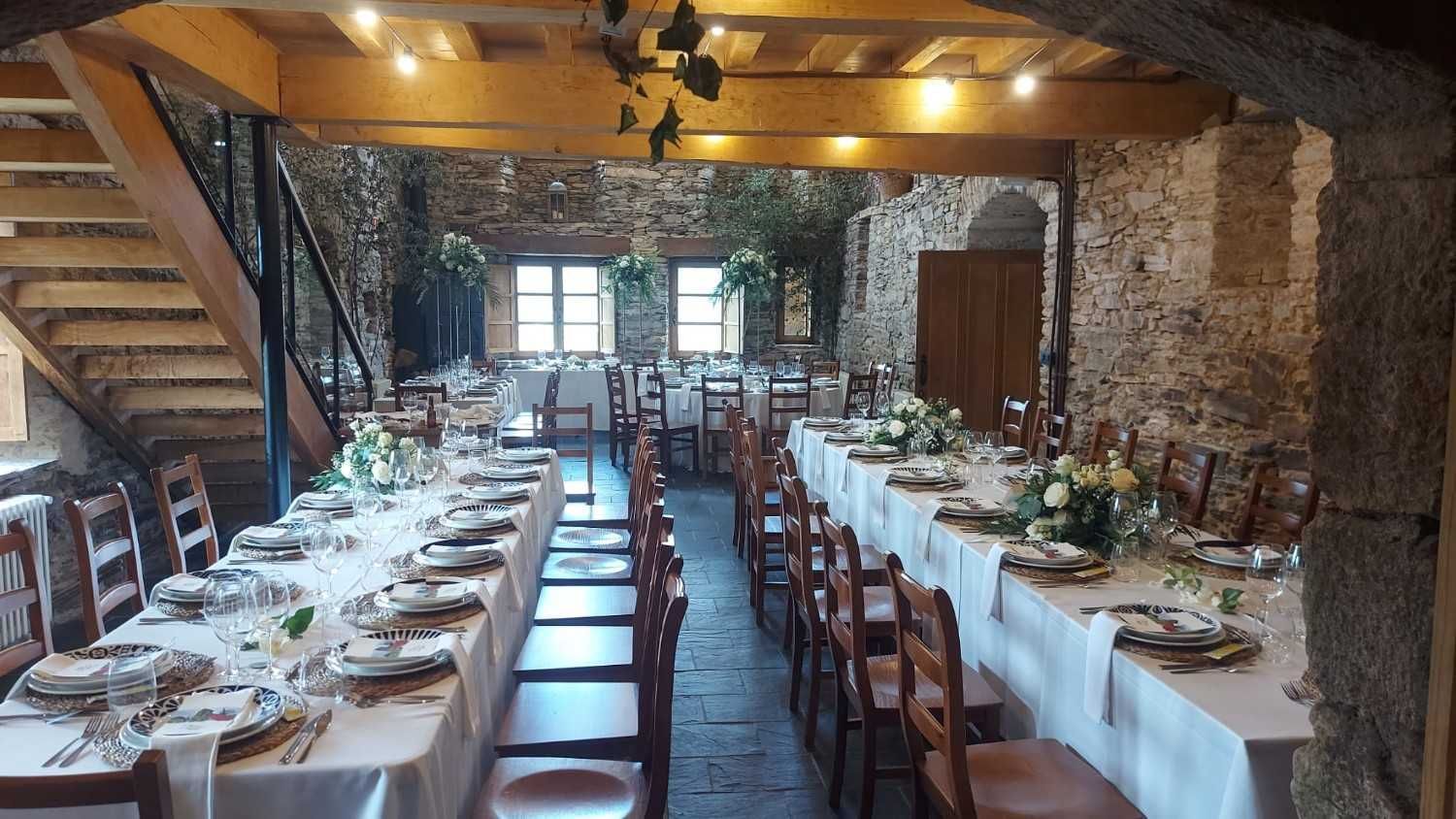 Mesas largas dispuestas para una recepción de boda en una sala con paredes de piedra y una escalera.