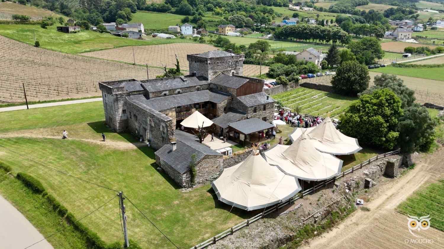Vista aérea de un castillo de piedra con tiendas de campaña instaladas en el terreno; campos y un pequeño pueblo al fondo.