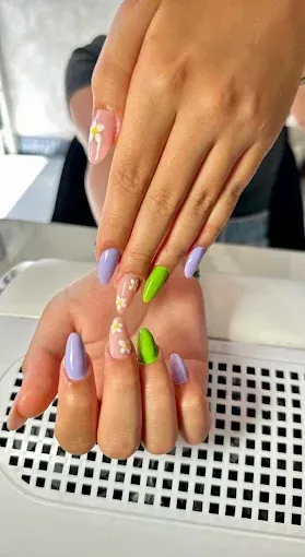 Manos con uñas en forma de almendra; diseños de color morado, verde y margaritas en una estación de manicura.