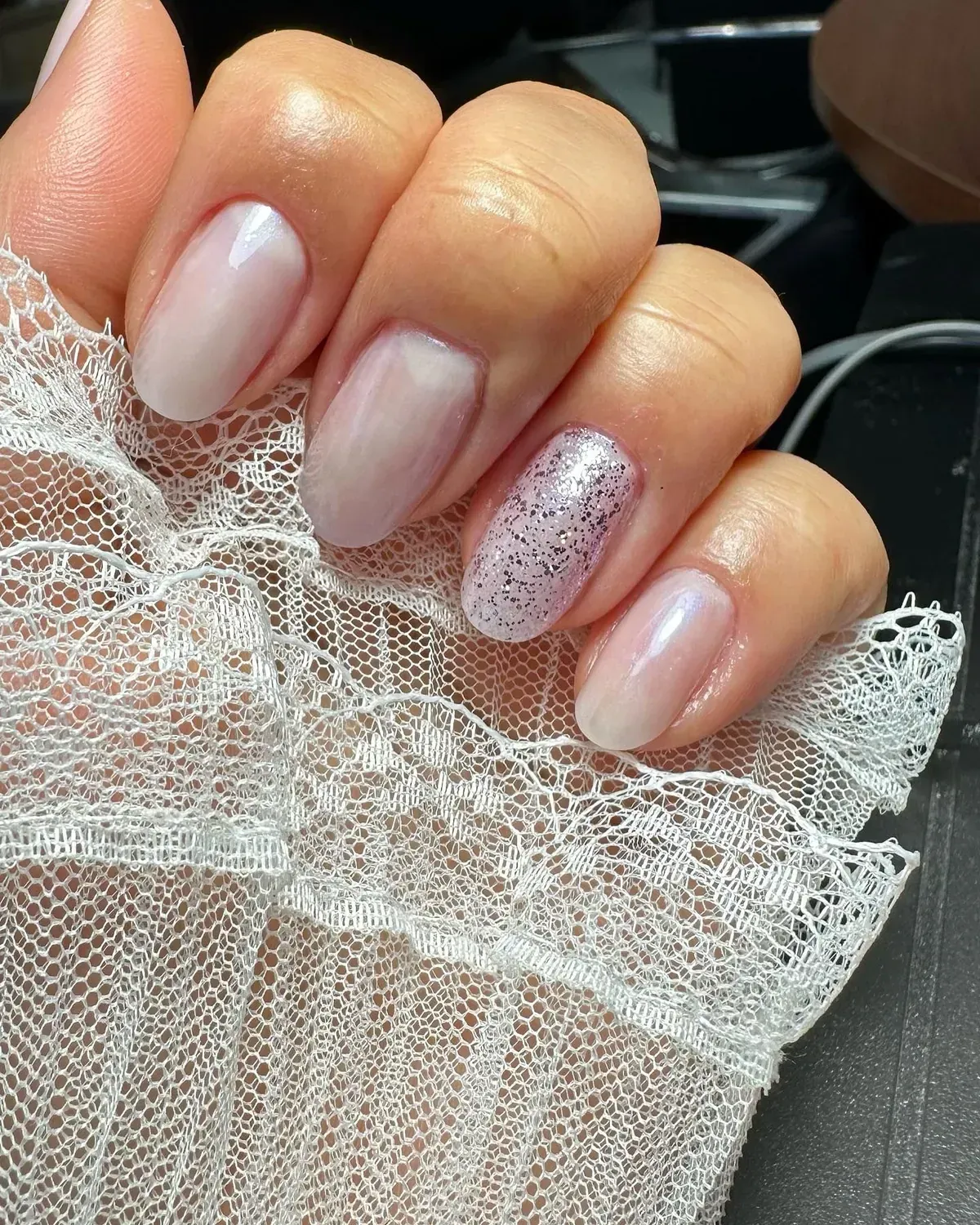 Primer plano de una mano con uñas ovaladas, mayormente rosa claro, una de ellas brillante. Apoyada sobre encaje blanco.