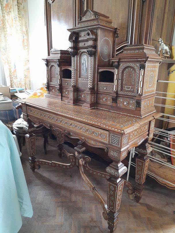 Bureau ancien en bois orné de sculptures dans une pièce baignée de soleil, près d’une fenêtre à rideaux.