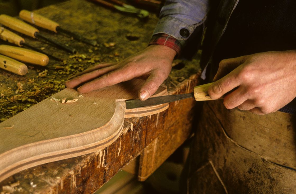 Des mains sculptent un objet en bois à l’aide d’un ciseau à bois sur un établi, dans un atelier