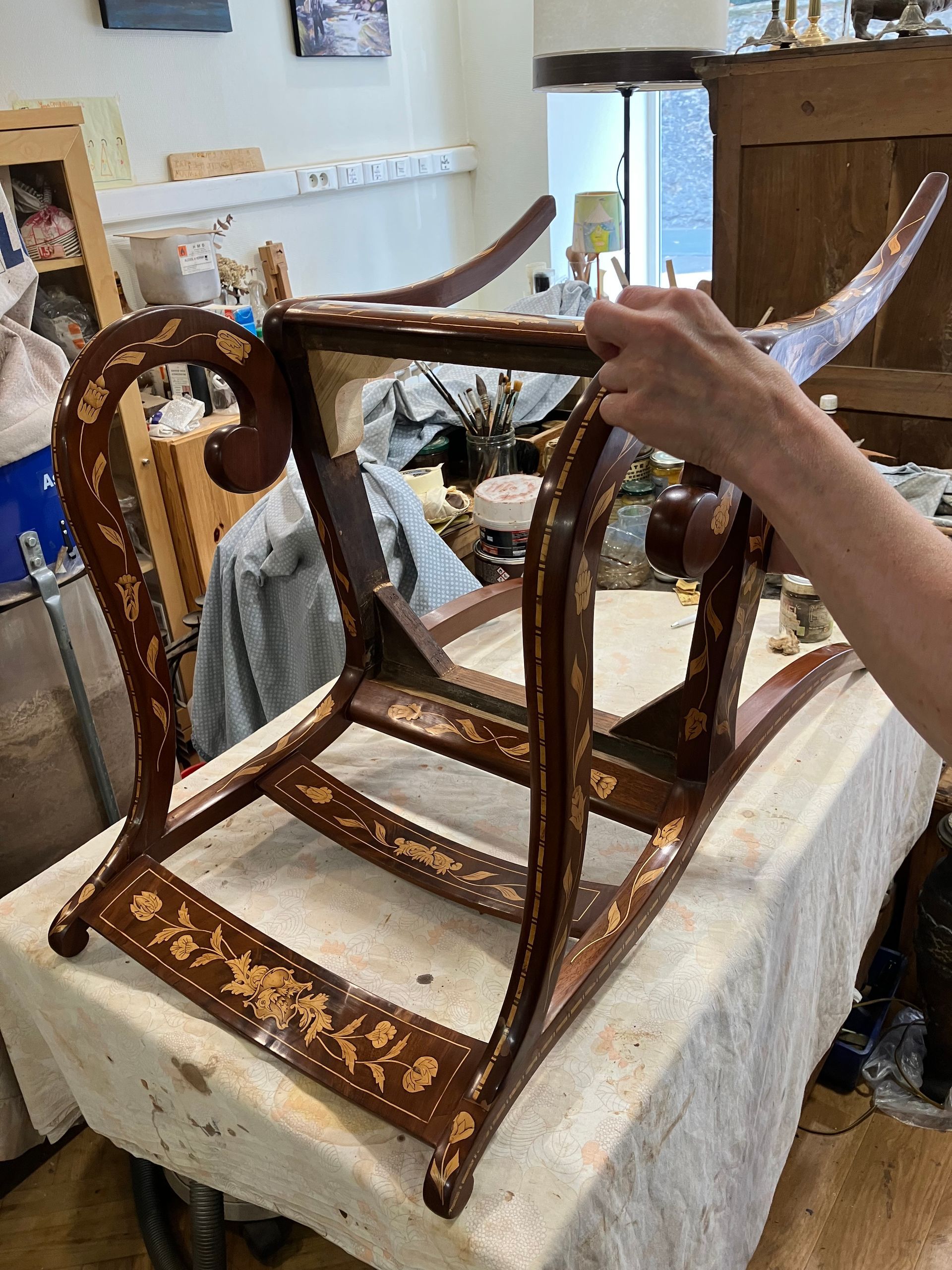 Une personne restaure le cadre sculpté d’une chaise en bois, posé sur un établi dans un atelier