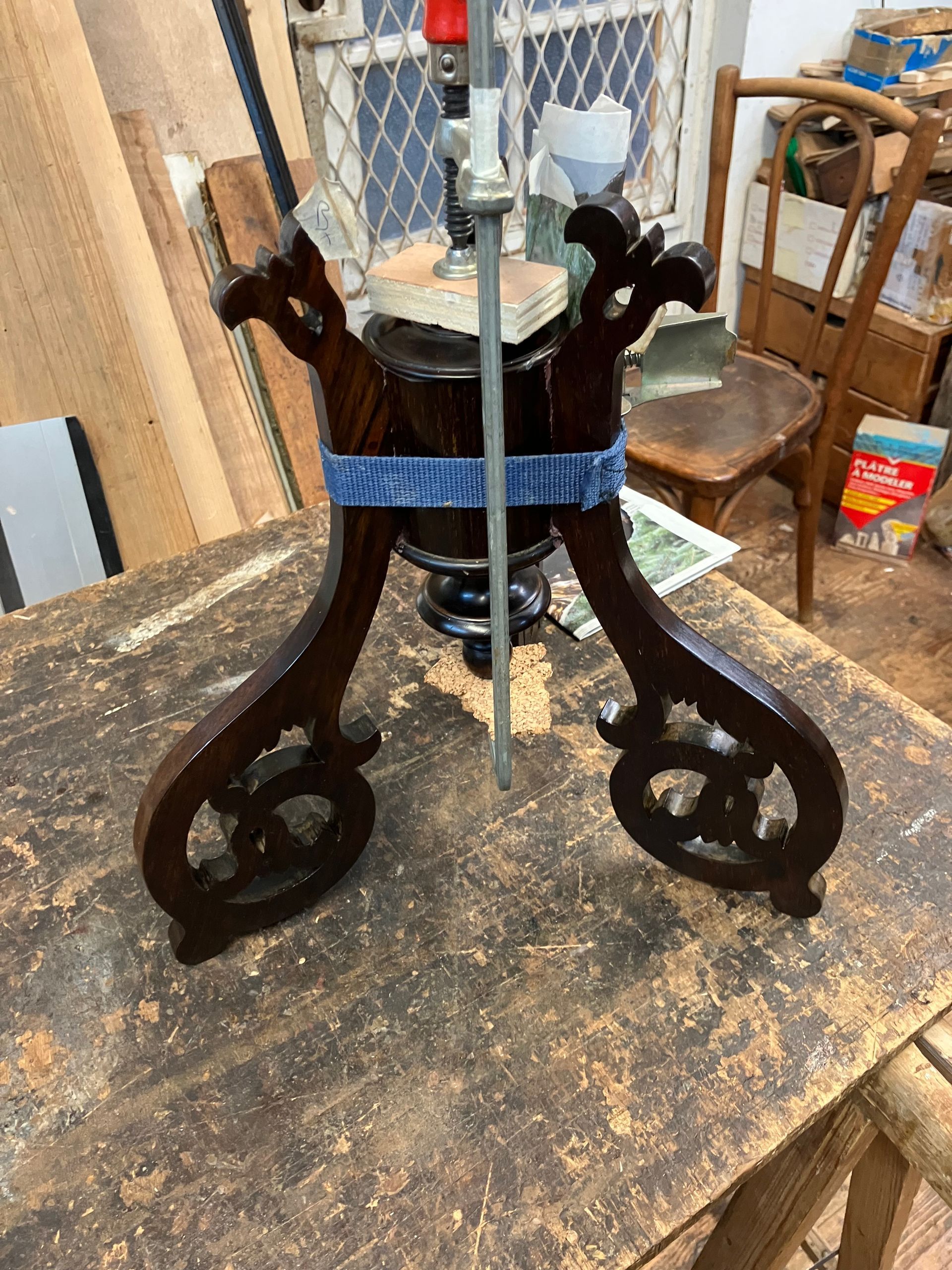 Support décoratif en bois foncé, aux pieds ornés et doté d’une sangle bleue, posé sur une table d’atelier
