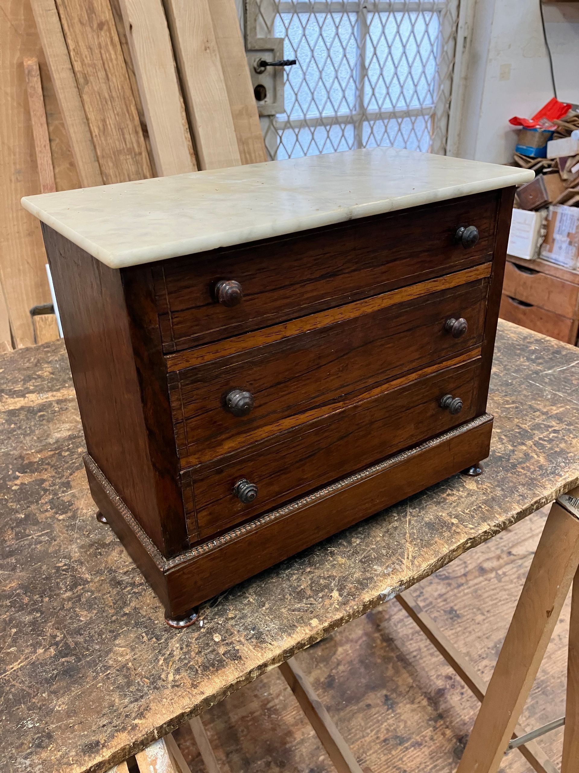 Petite commode en bois sombre avec trois tiroirs et un plateau en marbre clair, dans un atelier.