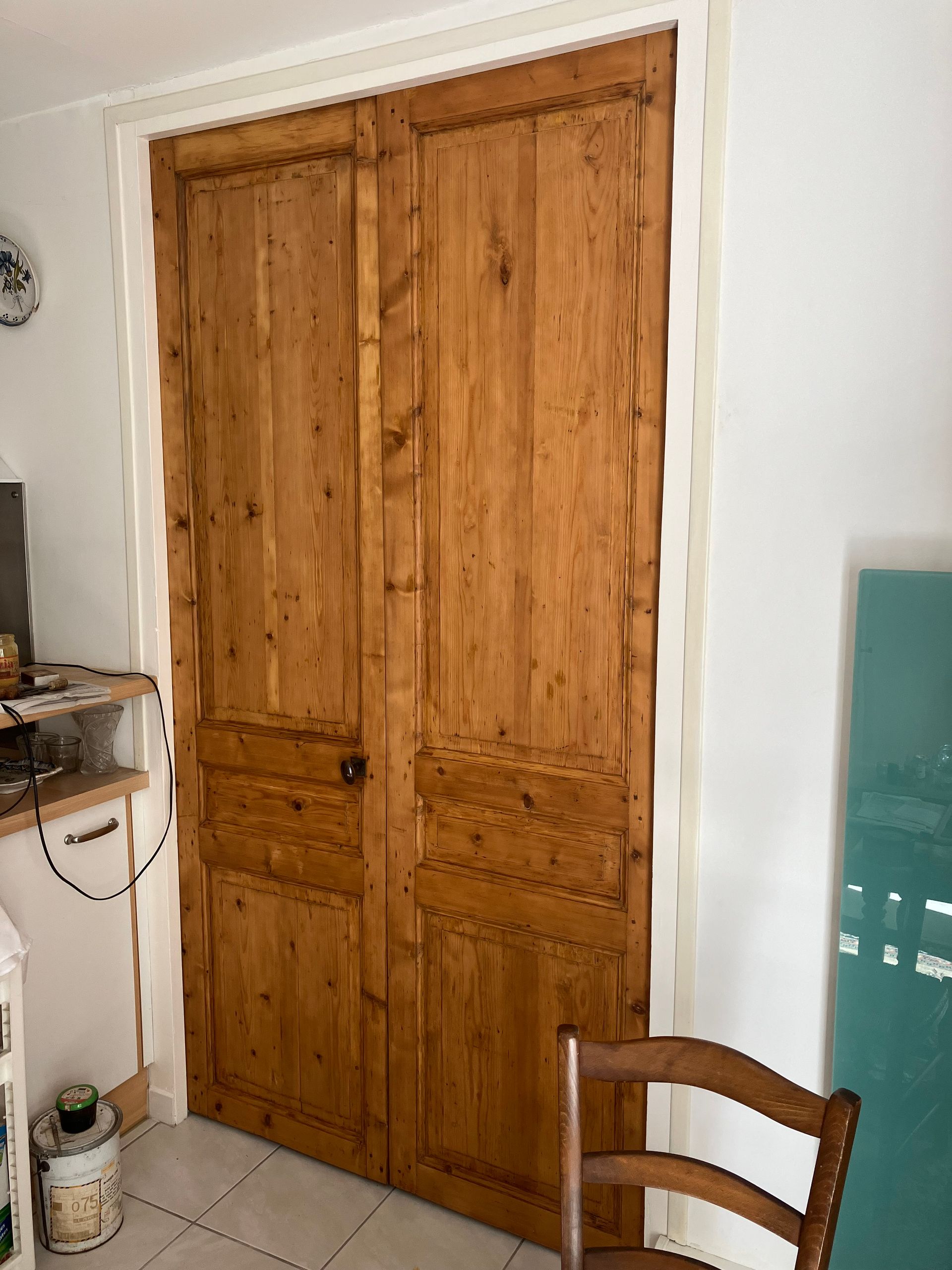 Armoire en bois à deux portes dans une pièce blanche, avec un meuble et une chaise bleu sarcelle partiellement visibles à proximité.
