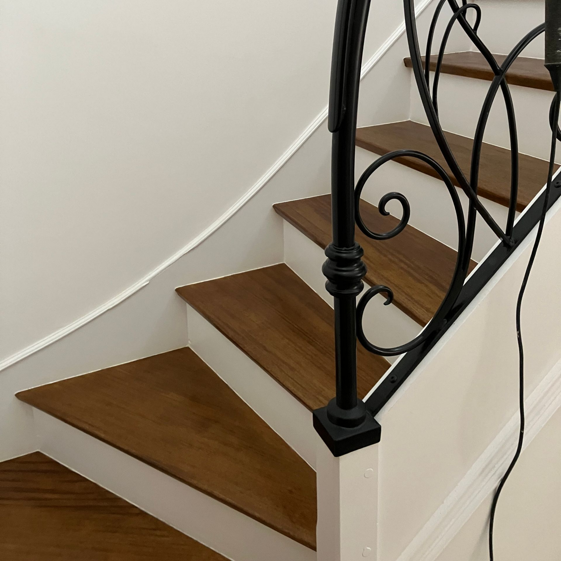 Escalier en bois avec des murs blancs, une rampe en fer forgé noir et un câble sur le côté droit.