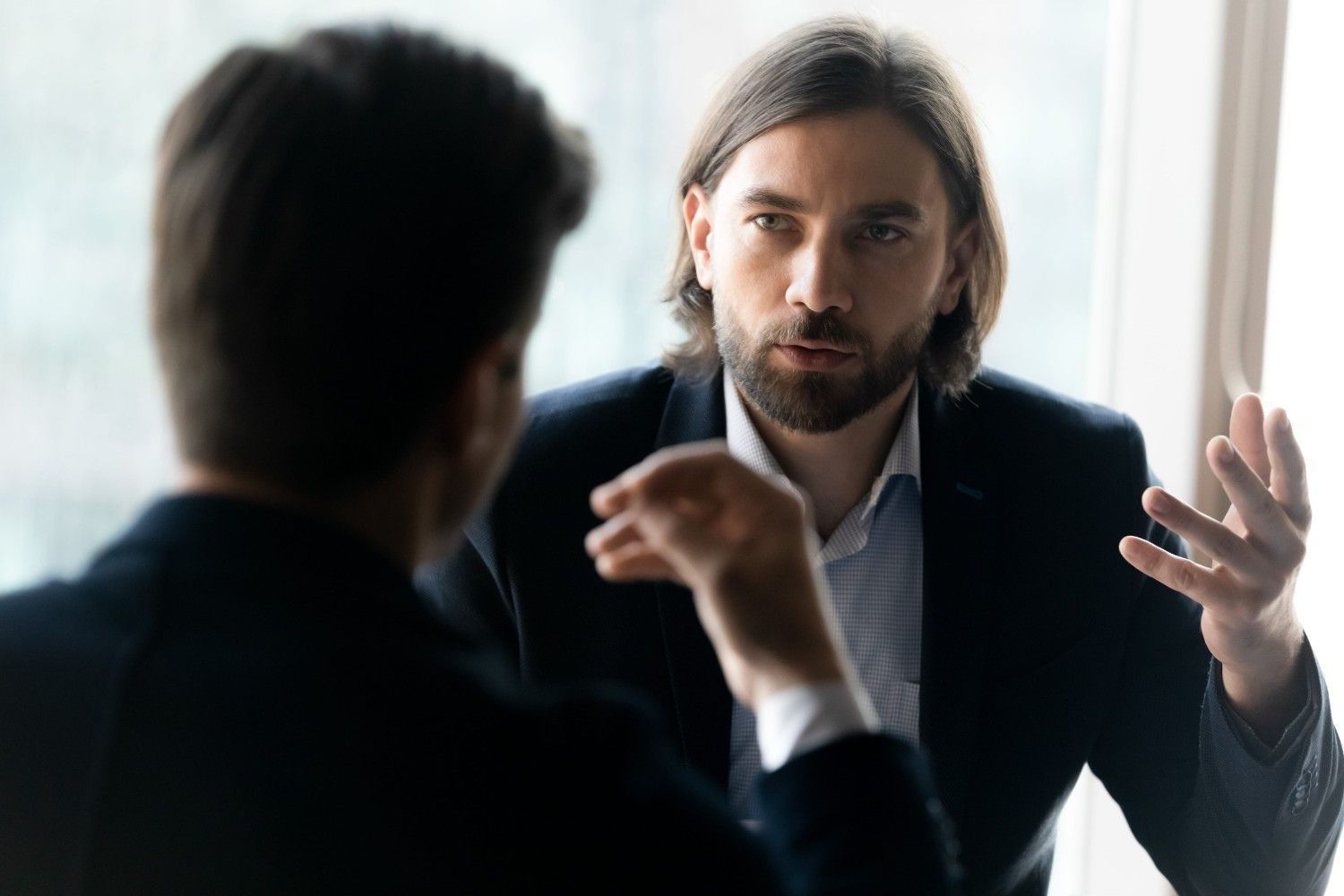 Dos personas en una oficina luminosa mantienen una conversación seria; una gesticula mientras la otra escucha.