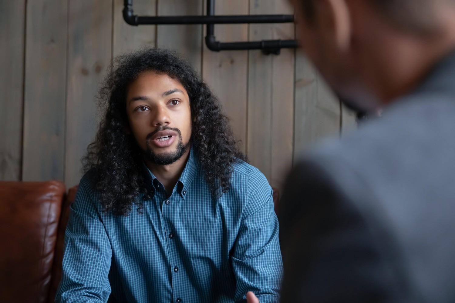 Un hombre con camisa azul habla en una habitación con paneles de madera mientras otra persona escucha en primer plano.