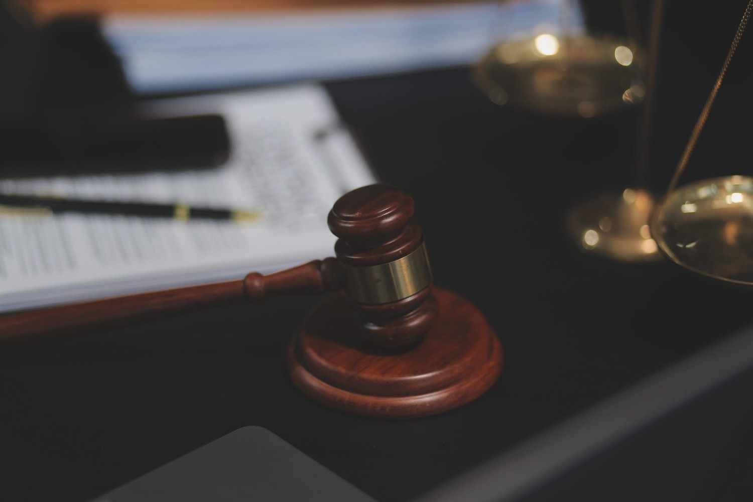 Mazo de juez de madera sobre un escritorio con documentos legales borrosos y balanzas al fondo.