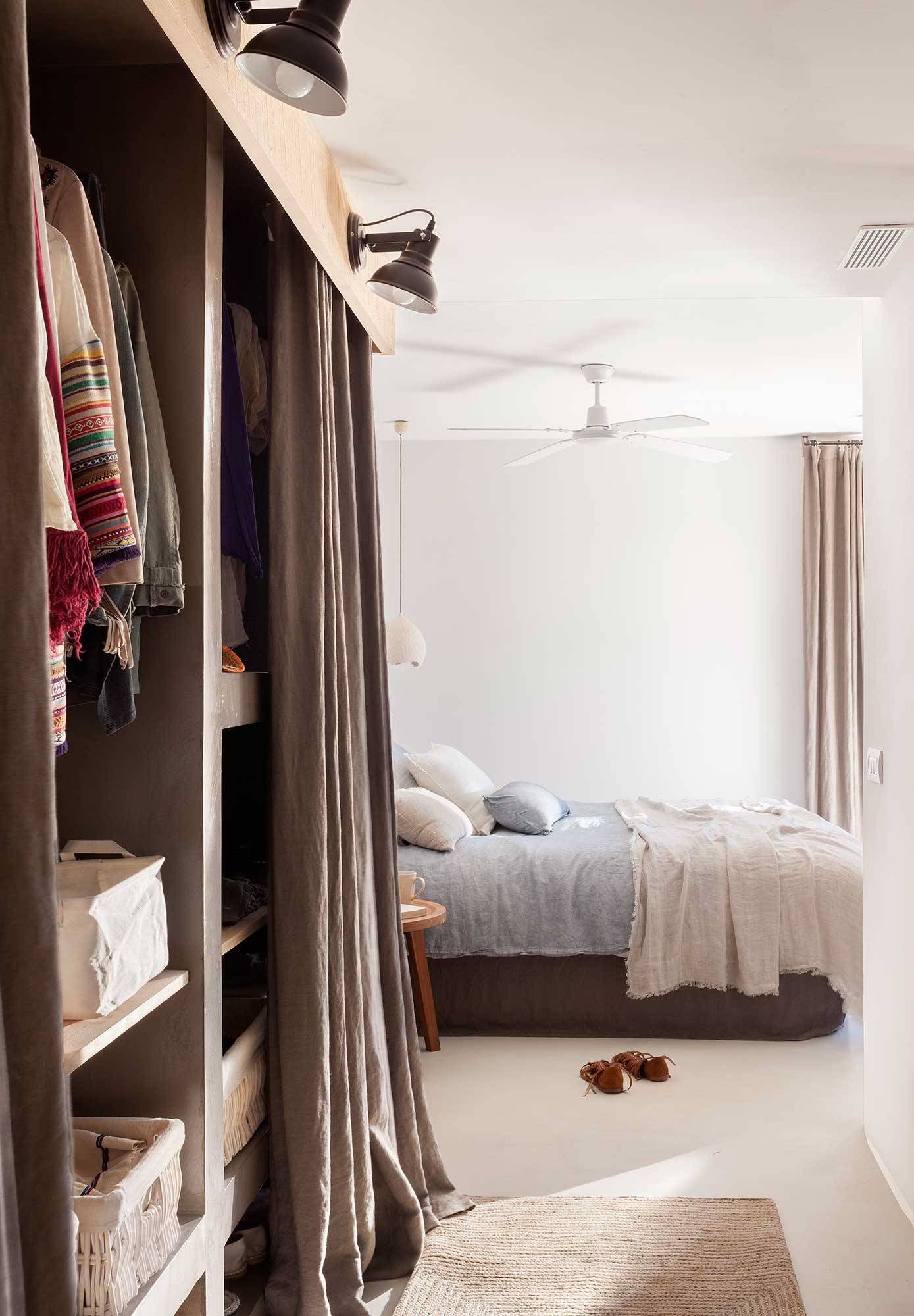 Dormitorio con armario abierto y cama a la vista. Cortinas grises, paredes blancas y detalles en madera marrón claro.