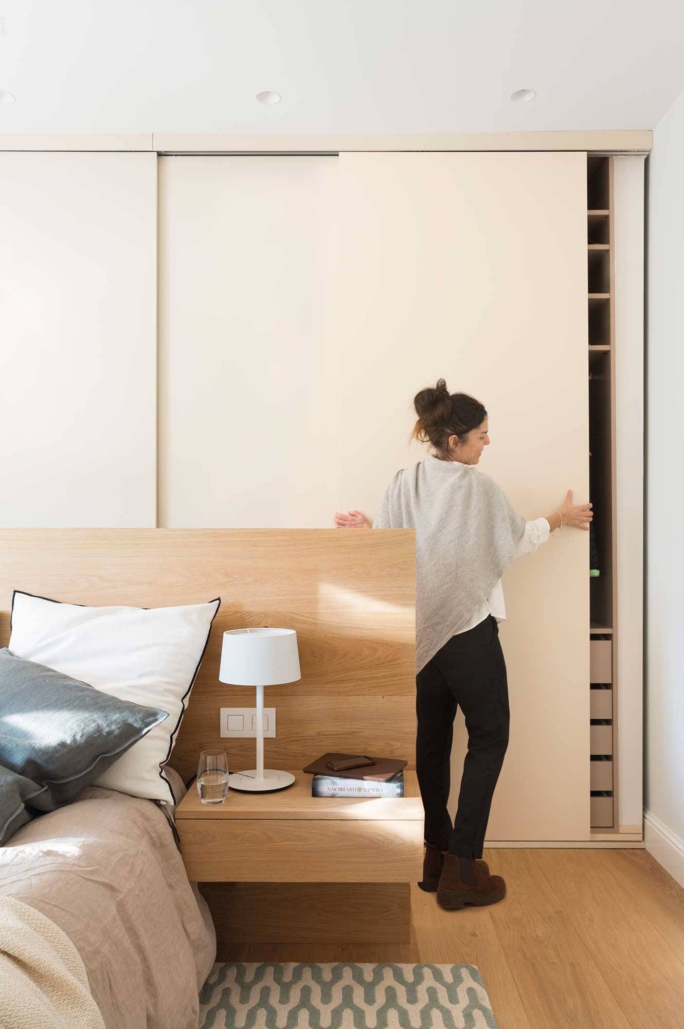Mujer abriendo la puerta corrediza de un armario en un dormitorio moderno con cabecero de madera y paredes de colores claros.