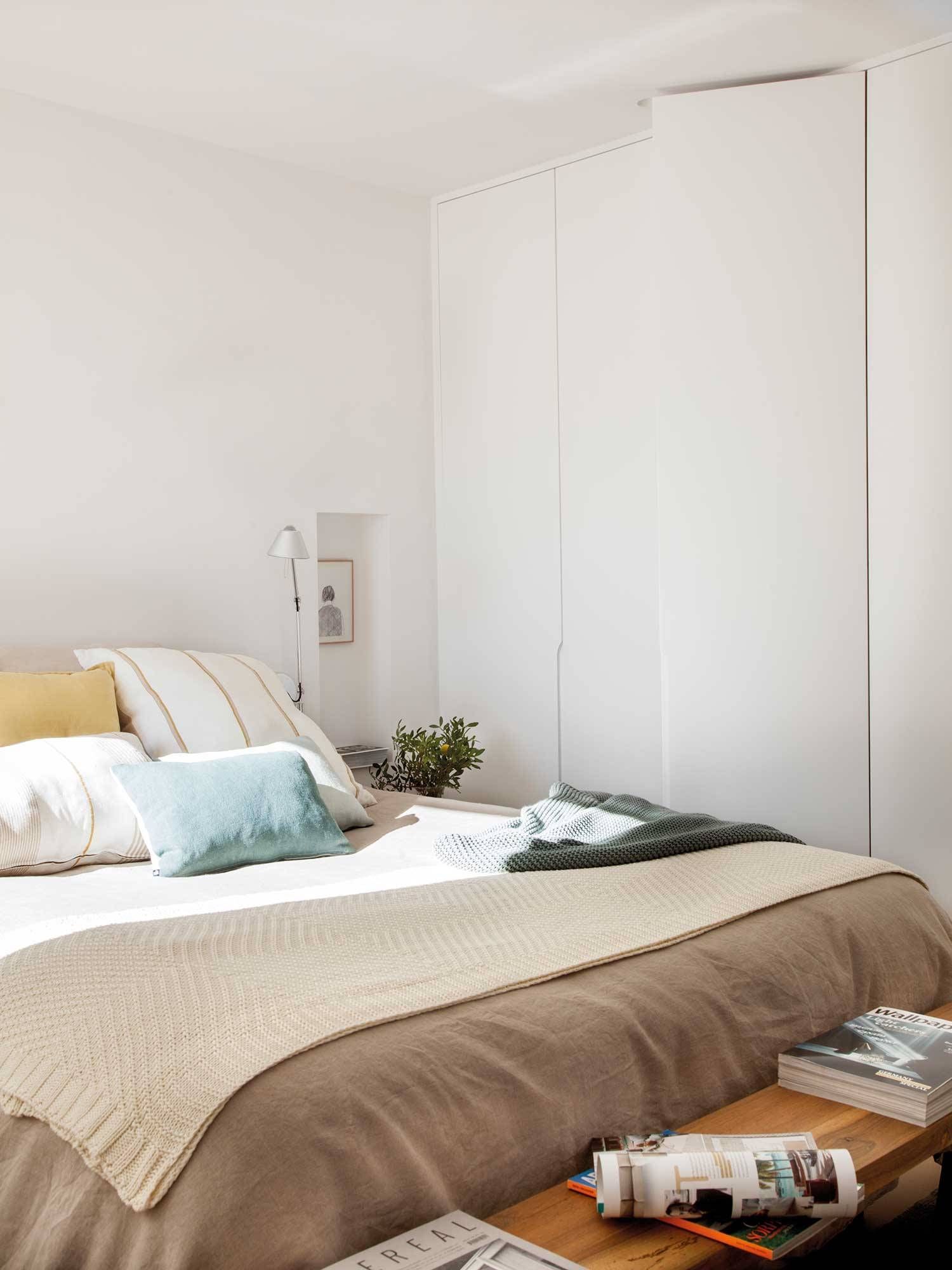 Dormitorio con paredes blancas, cama grande con ropa de cama neutra, almohadas y un banco de madera con libros.