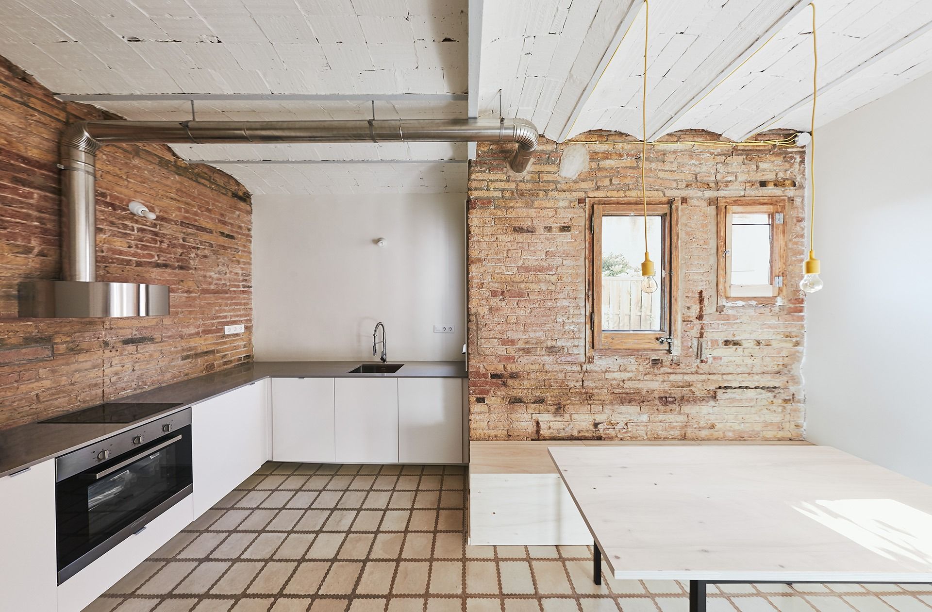 Cocina moderna con paredes de ladrillo a la vista, gabinetes blancos y una mesa de madera sencilla.