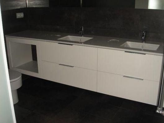 Tocador blanco de dos lavabos con encimera gris sobre pared y suelo de azulejos oscuros. Estante abierto a la izquierda.