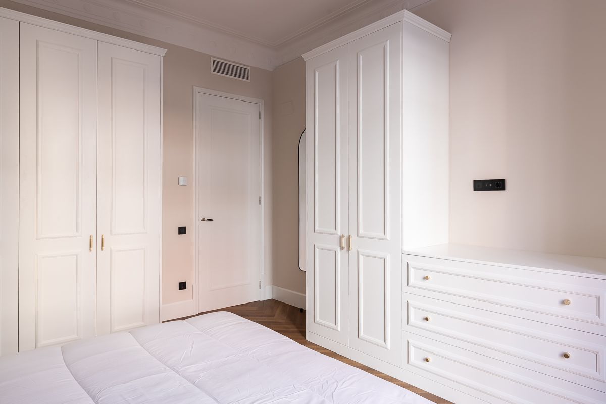 Dormitorio blanco con tres armarios, una puerta y una cómoda.