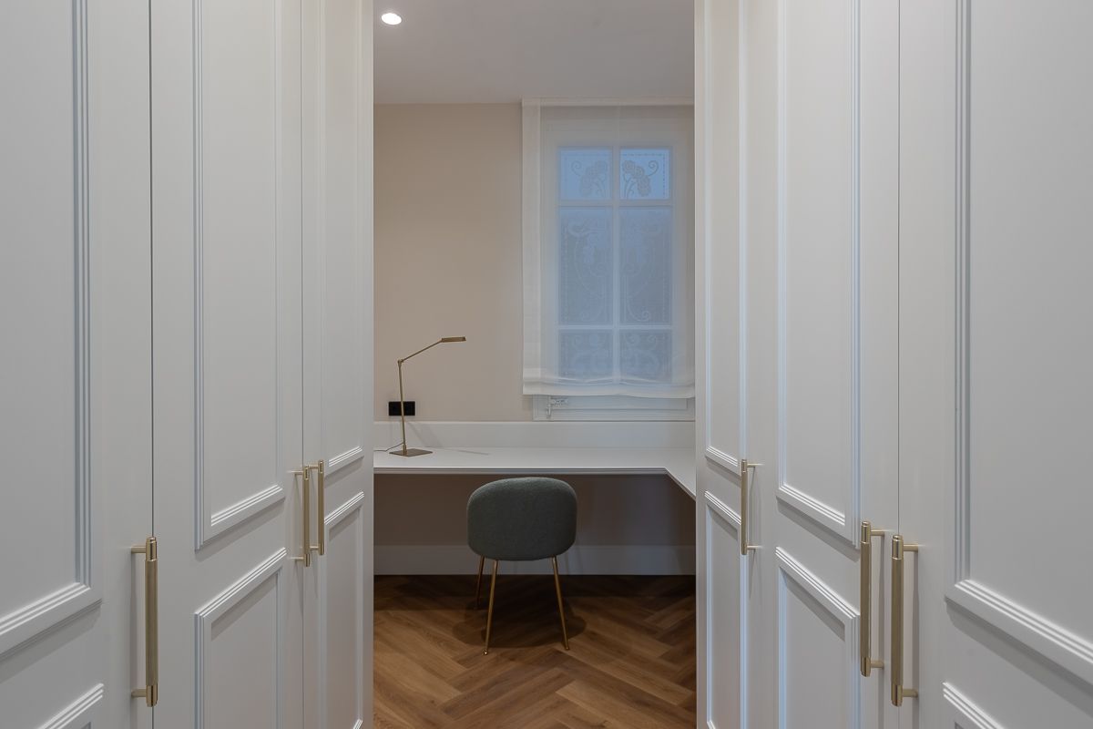 Un estrecho pasillo blanco conduce a un escritorio con una silla junto a una ventana. Suelos de madera y herrajes dorados.