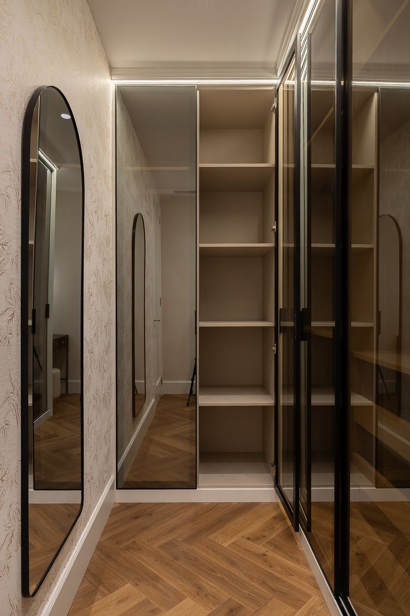 Vestidor con estantes y puertas de vidrio, piso de madera y grandes espejos arqueados.
