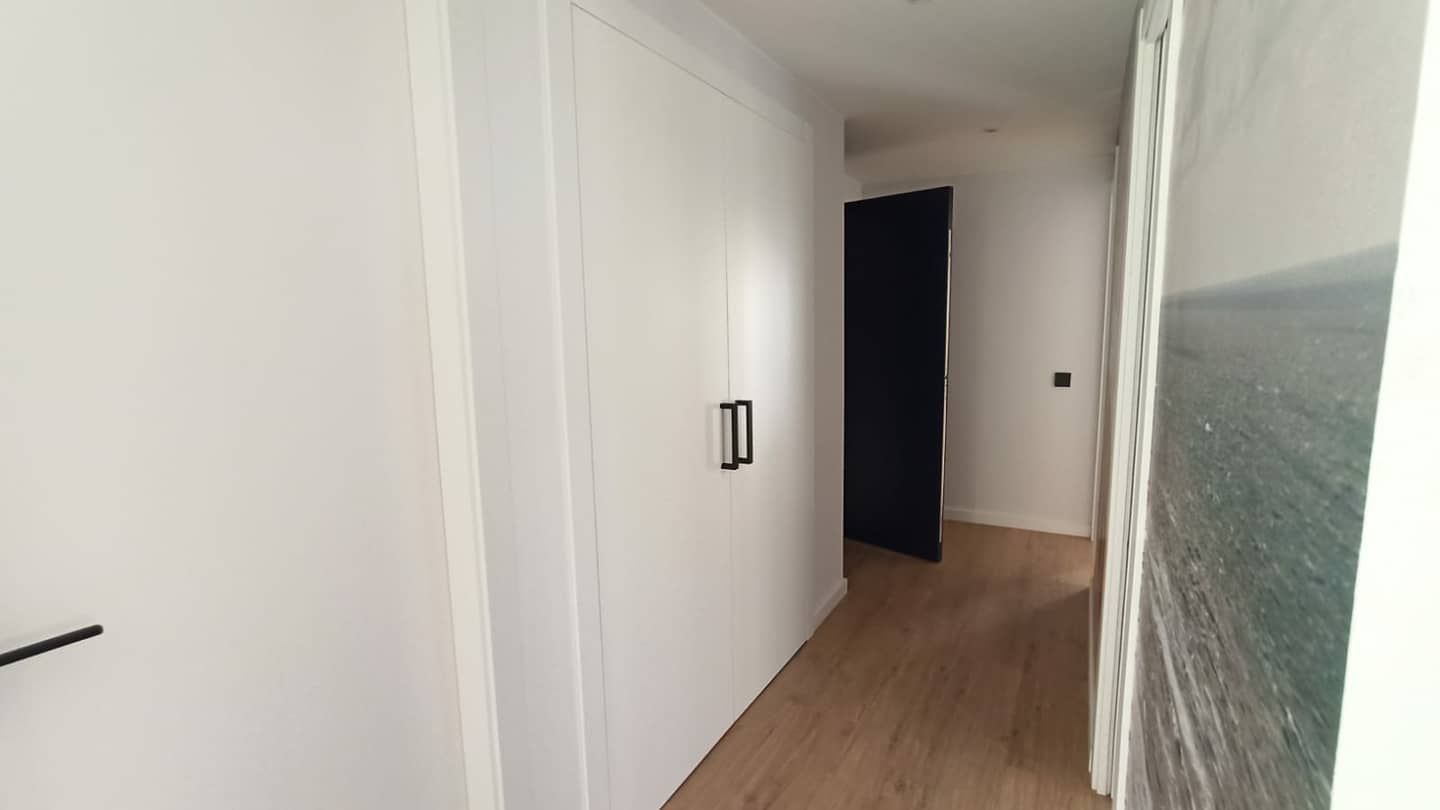 Pasillo blanco con puertas de armario, manija negra, puerta negra abierta al final y obra de arte en la pared.