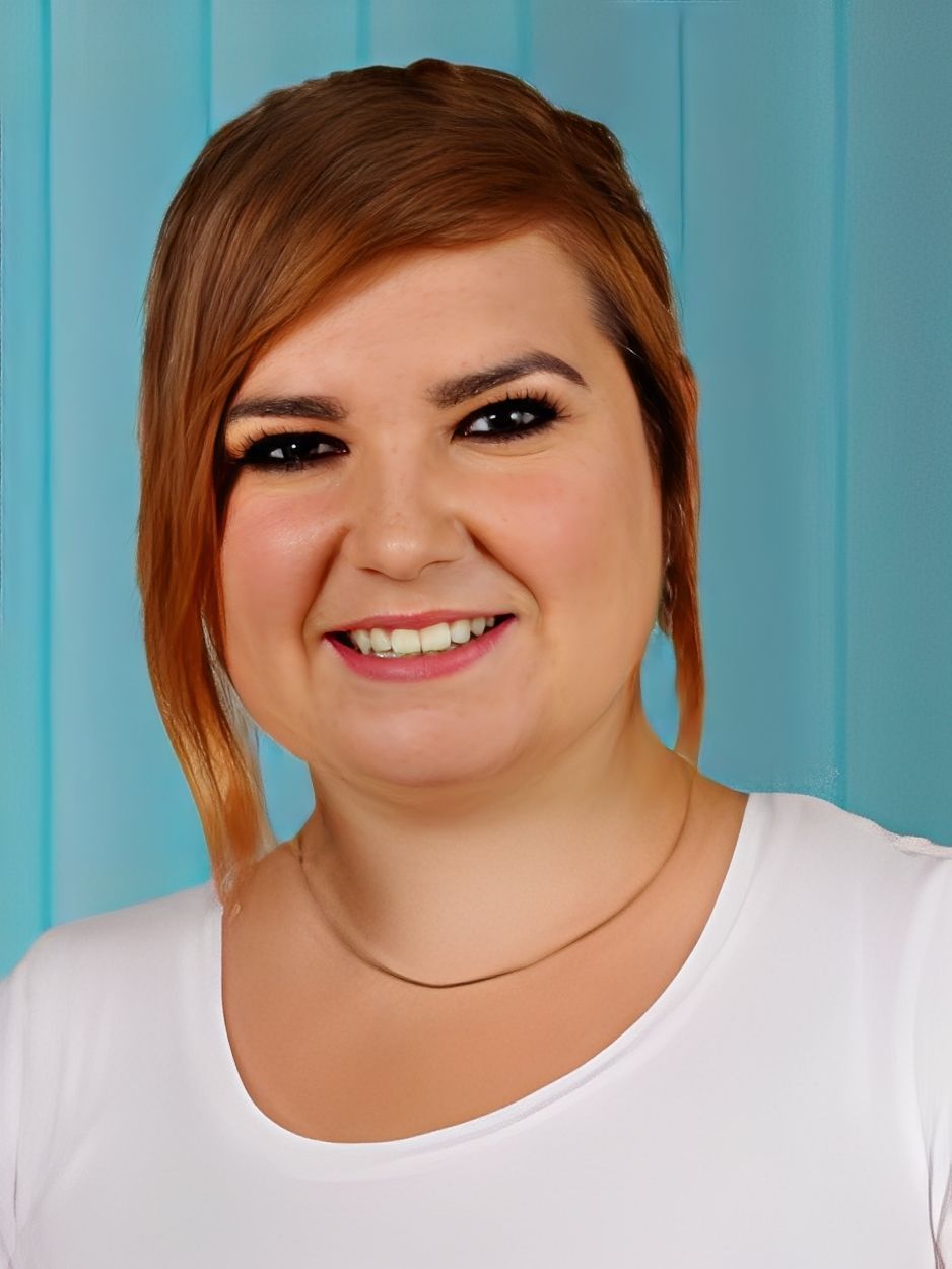 Frau mit roten Haaren lächelt in die Kamera, trägt ein weißes Hemd vor einem blauen Hintergrund.