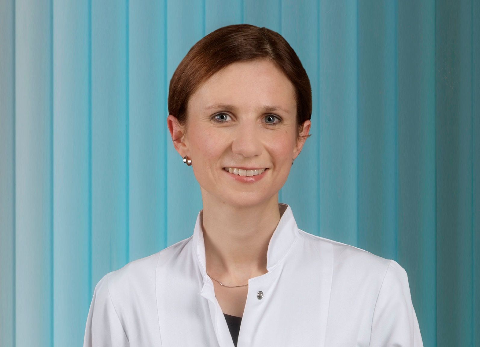Frau im weißen Laborkittel lächelt und steht vor blau gestreiftem Hintergrund.